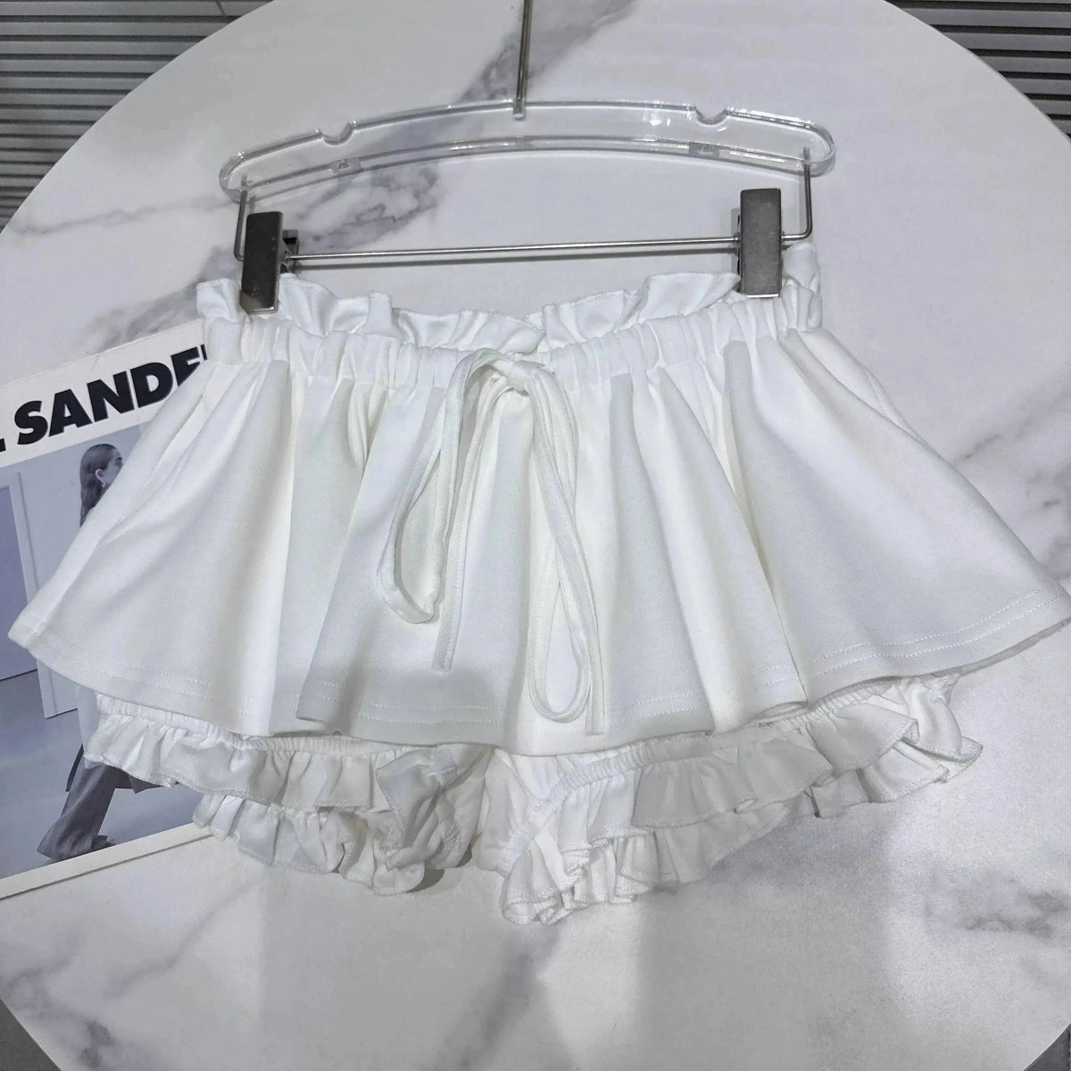 A pair of white frilly shorts with an elasticized ruffled waistband and a drawstring tie, shown hanging on a hanger against a white marble tabletop.