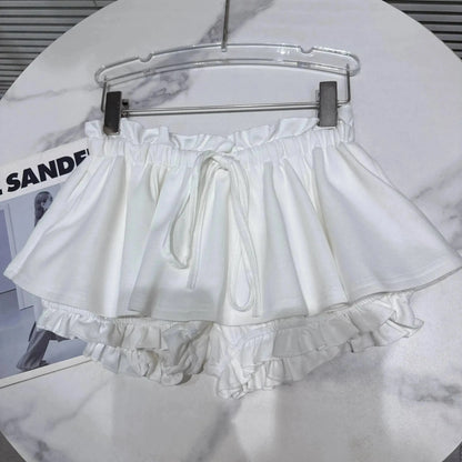 A pair of white frilly shorts with an elasticized ruffled waistband and a drawstring tie, shown hanging on a hanger against a white marble tabletop.