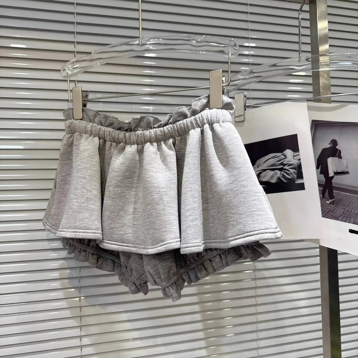 A pair of light gray ruffled sweat shorts hanging on a clear hanger against a white slatted blind background.