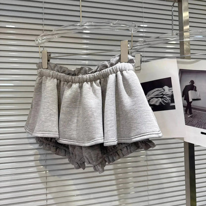A pair of light gray ruffled sweat shorts hanging on a clear hanger against a white slatted blind background.