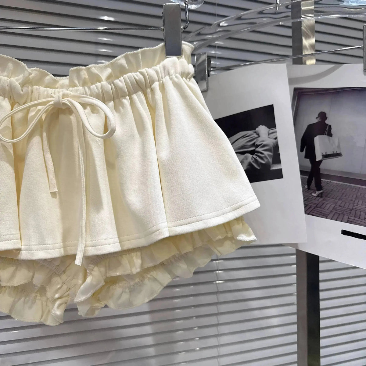 Cream-colored ruffled shorts with an elasticated drawstring waistband hanging on a display rack next to hanging photography prints.