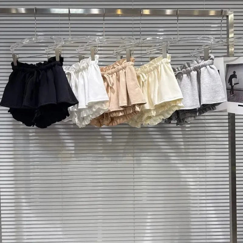 Five pairs of ruffled elastic-waist shorts in black, white, tan, cream, and grey hanging on a metal rack against a white slatted wall.