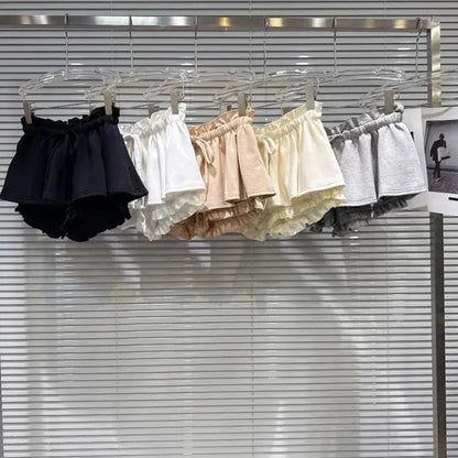 Five pairs of ruffled elastic-waist shorts in black, white, tan, cream, and grey hanging on a metal rack against a white slatted wall.
