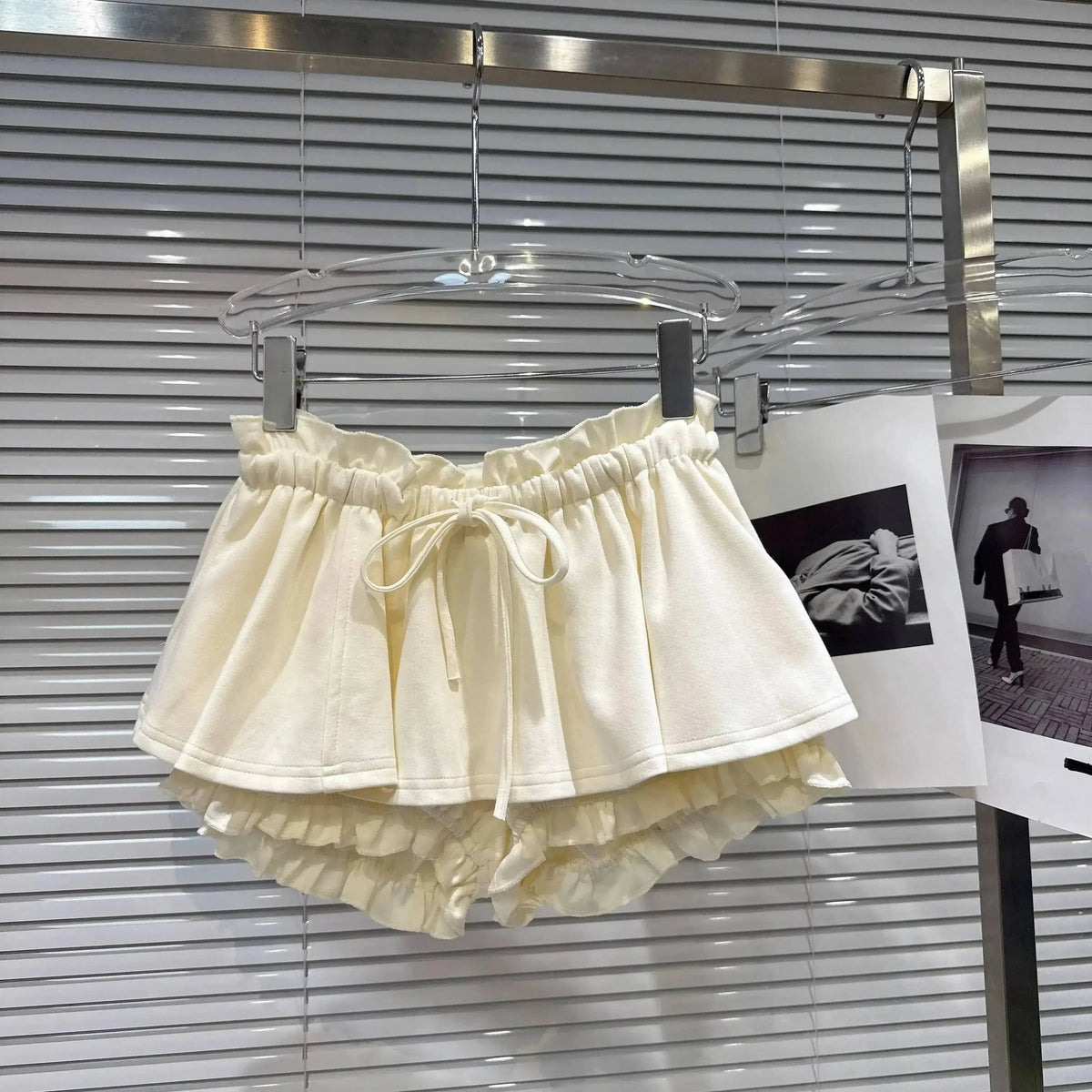 A pair of cream-colored ruffled shorts with an elastic drawstring waistband hangs on a clear hanger against a white horizontal blind background.