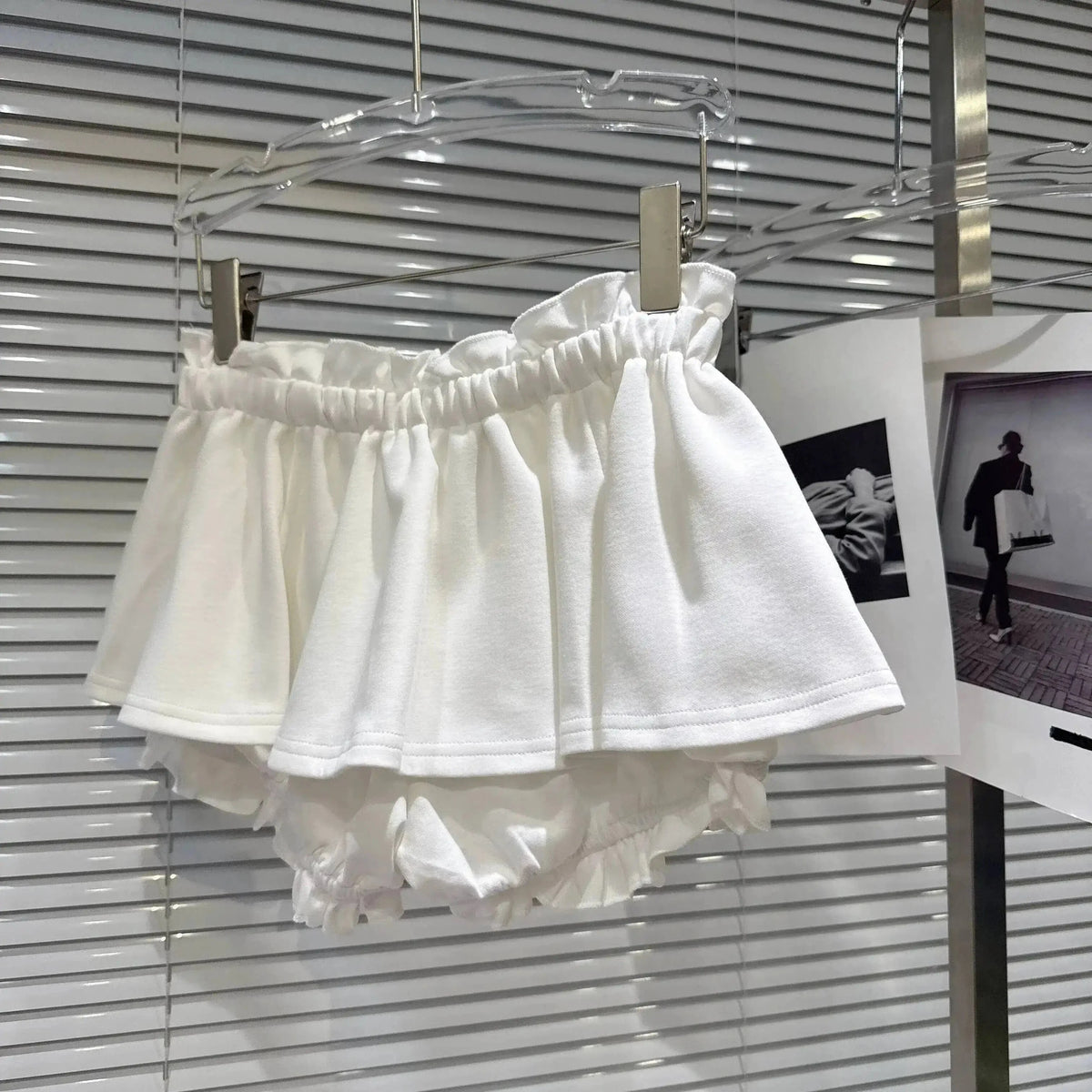 A pair of white frilled bloomers hanging from a clear hanger against a white slatted background.