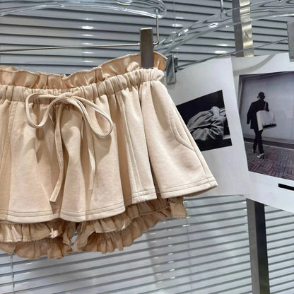 Beige tiered ruffle shorts with an elastic paperbag waist and a matching drawstring tie, hanging on a clothing rack in front of displayed photographs.