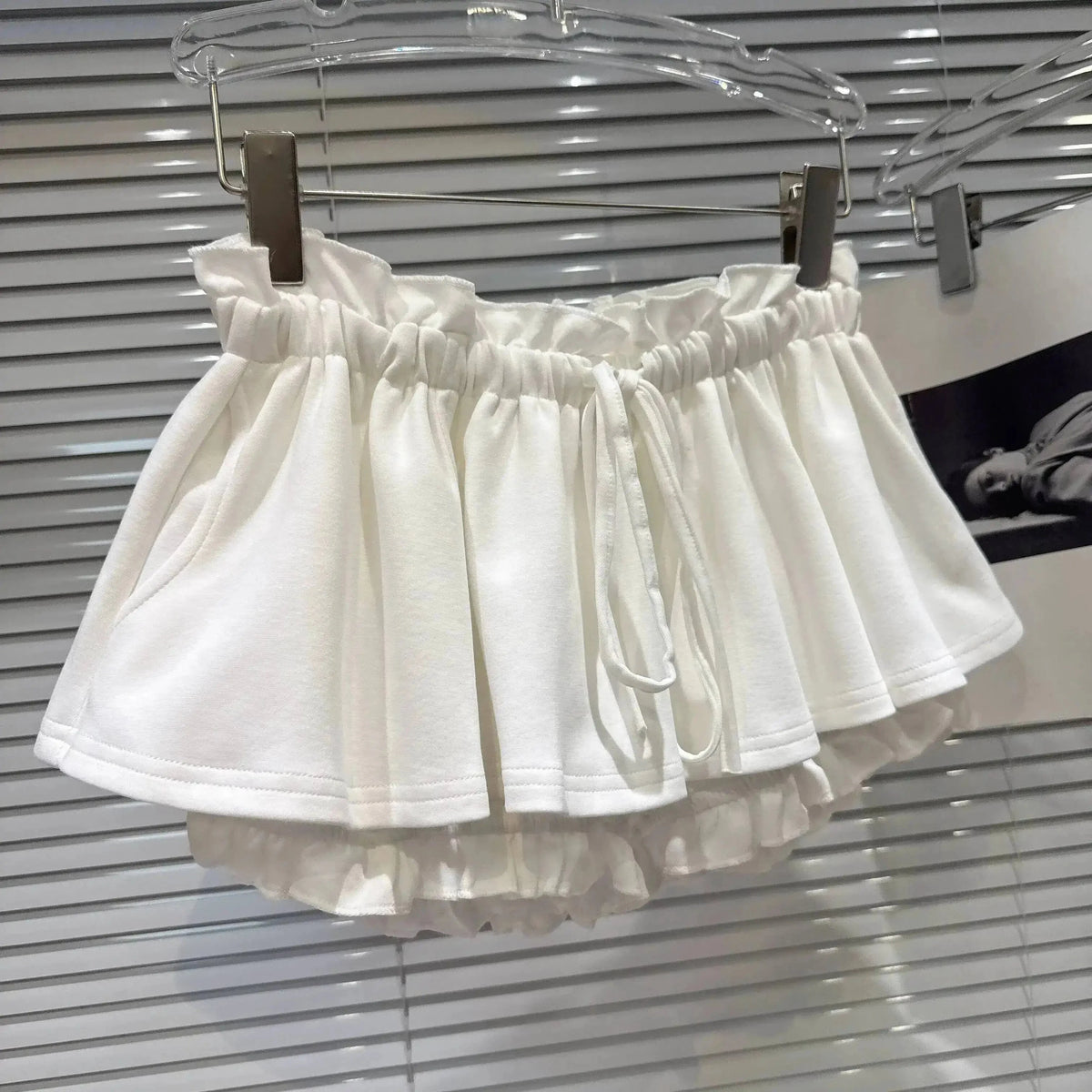 A white tiered mini skirt with an elasticated, ruffled waistband and a decorative drawstring hanging from a silver hanger against a grey slatted background.