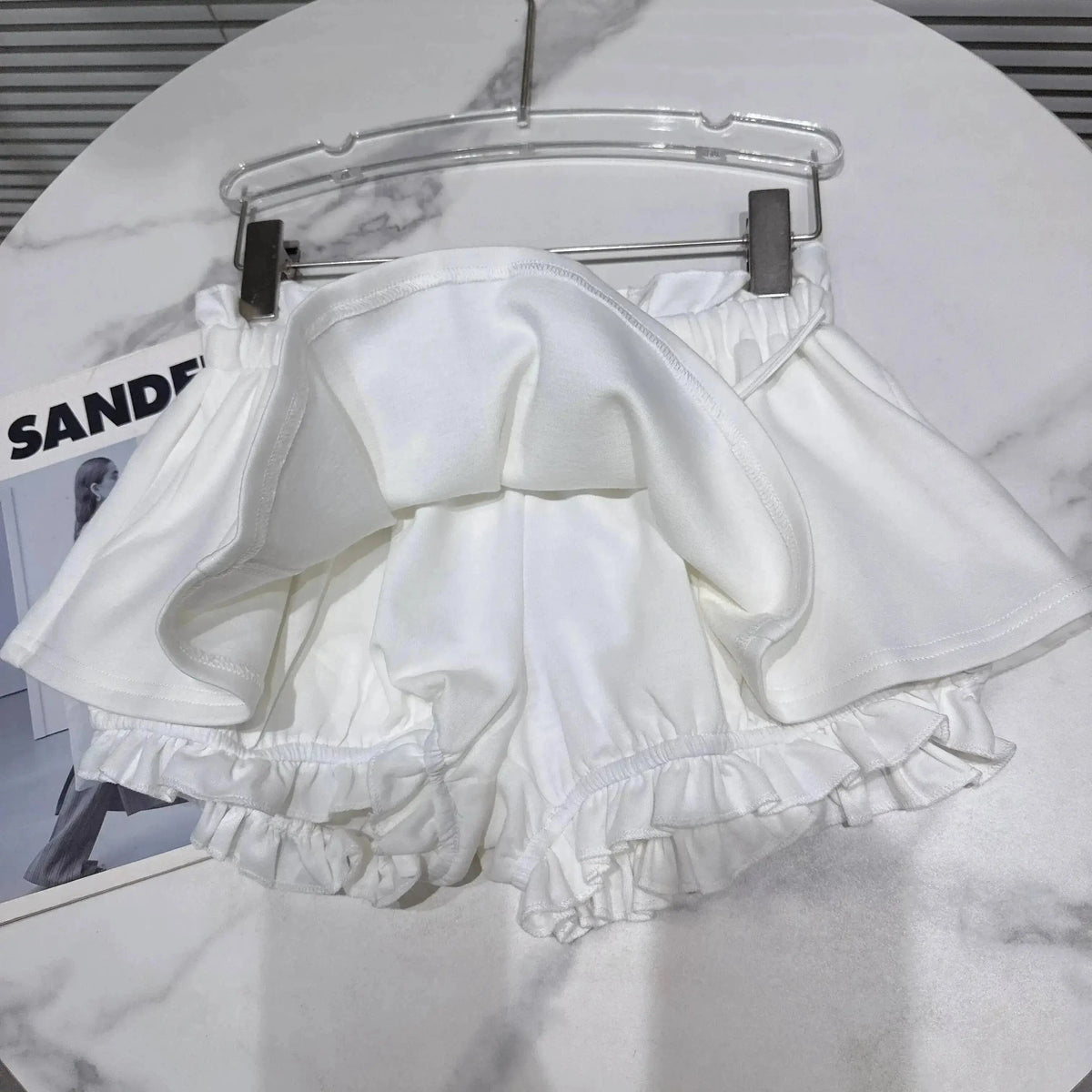 A white skort hangs on a clear plastic hanger, featuring a ruffled hem and a pleated overlay design.