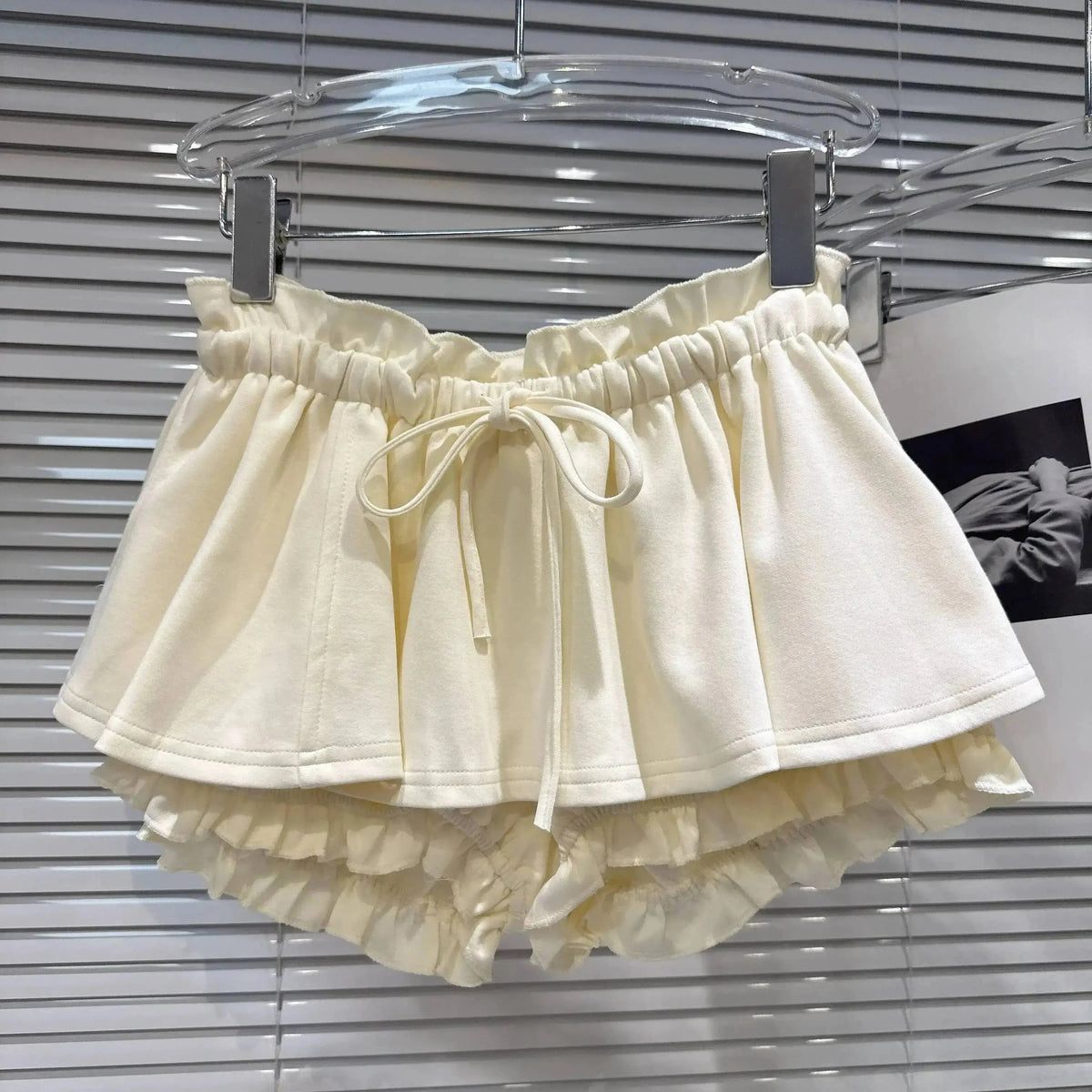 Cream-colored ruffled shorts with an elasticated drawstring waistband hanging on a clear hanger in front of white horizontal blinds.