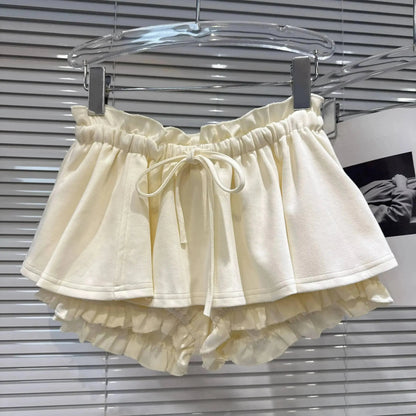 Cream-colored ruffled shorts with an elasticated drawstring waistband hanging on a clear hanger in front of white horizontal blinds.
