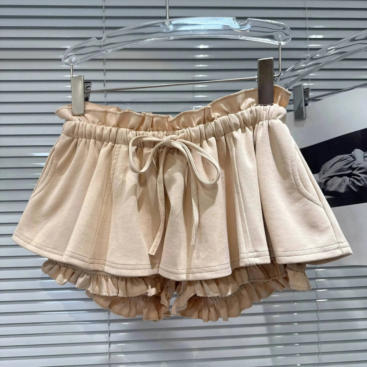 A beige skirt-style pair of shorts with an elasticated waist, drawstring tie, and a layered ruffled hem hanging on a hanger.