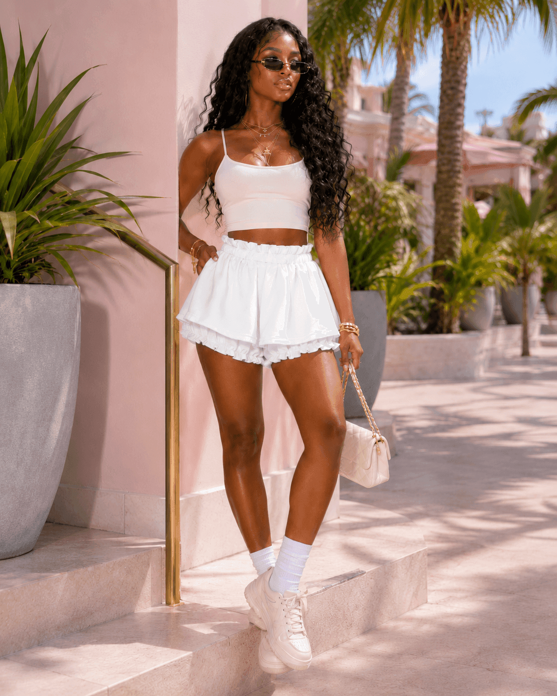 A woman wearing a white camisole top and matching ruffled high-waisted shorts stands outdoors near a pale pink wall with palm trees in the background.