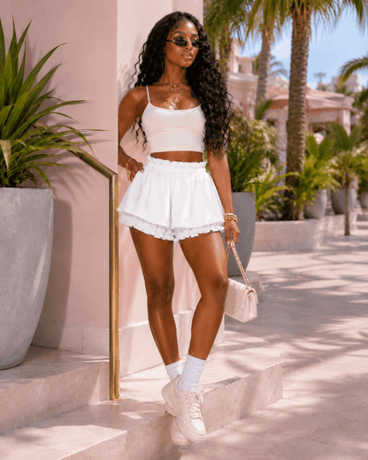 A woman wearing a white camisole top and matching ruffled high-waisted shorts stands outdoors near a pale pink wall with palm trees in the background.
