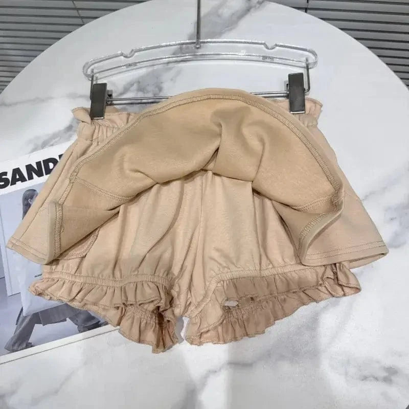 A beige skort hangs from a hanger on a marble surface, featuring a ruffled hem layer over built-in shorts with an elasticized waistband.