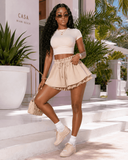 Beige high-waisted tiered ruffle shorts with a drawstring waist worn with a fitted short-sleeve crop top, white crew socks, and tan sneakers in a sunny outdoor setting.