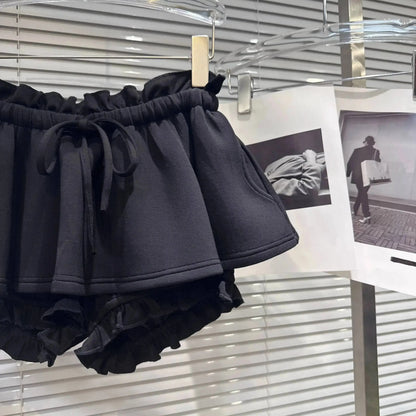 Black ruffled shorts with an elasticated drawstring waistband hanging on a display rack in front of white blinds.