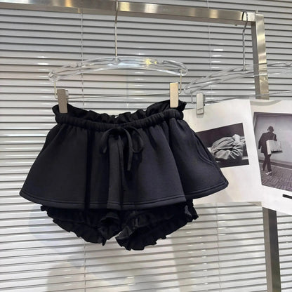 Black ruffled shorts with an elastic paperbag waist and a matching drawstring tie, hanging from a transparent hanger against a horizontal slat blind.