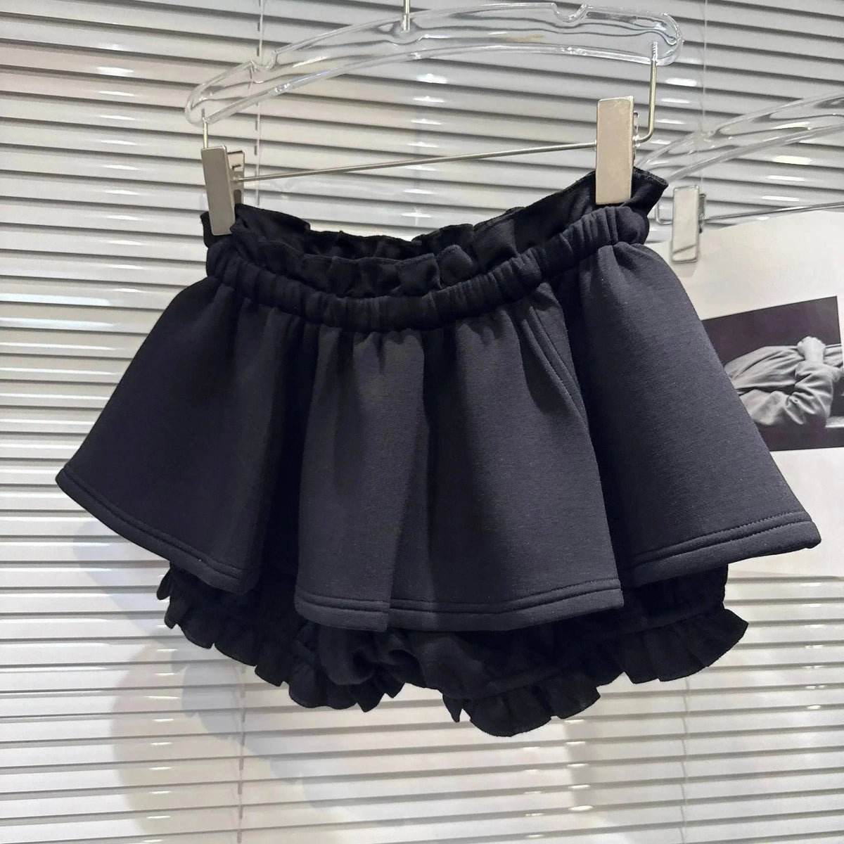 A black ruffled mini skirt with an elastic waistband hangs from a transparent hanger against a white slatted background.