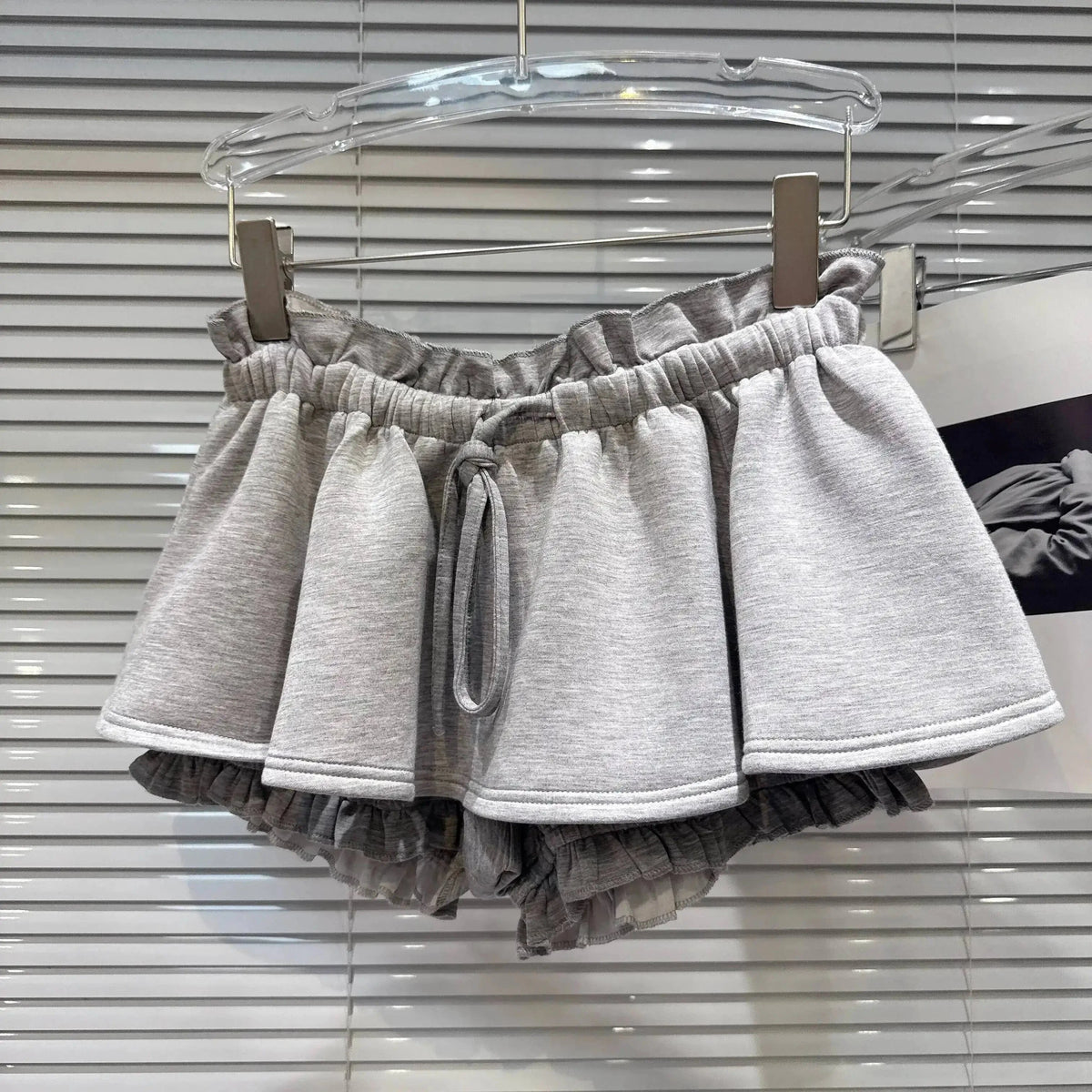 Gray ruffled shorts with an elastic paperbag waist and a matching drawstring tie, hanging against white window blinds.