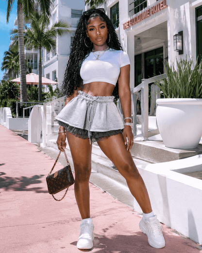 A woman wearing a white cropped t-shirt and grey layered ruffled shorts with a drawstring waist stands outside on a pink walkway next to a white building.