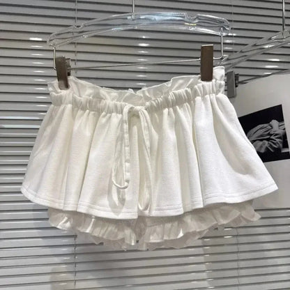 A white, tiered ruffled mini skirt or shorts with an elasticated waistband and a drawstring closure, displayed on a hanger against a slatted blind background.