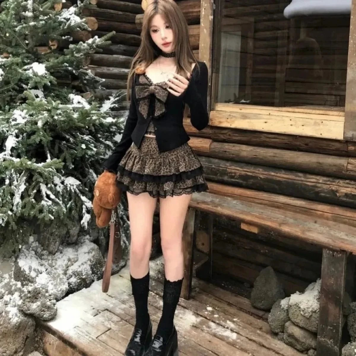 A young woman wears a black long-sleeved cardigan over a leopard-print bustier top with a matching tiered mini skirt and black lace-trimmed knee-high socks, standing outdoors in front of a wooden cabin in the snow.
