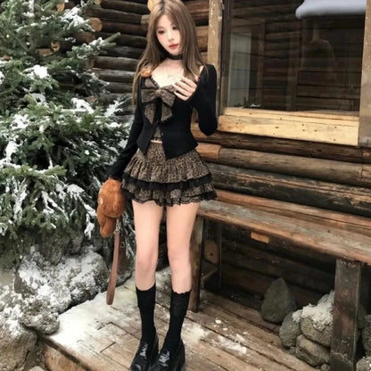 A young woman wears a black long-sleeved cardigan over a leopard-print bustier top with a matching tiered mini skirt and black lace-trimmed knee-high socks, standing outdoors in front of a wooden cabin in the snow.