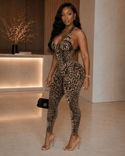 A woman wears a sleeveless, leopard-print jumpsuit with a halter neckline and cut-out sides, paired with gold strappy heels and a small black handbag in a modern interior setting.