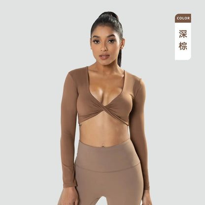 A woman wears a long-sleeved, deep-brown cropped top featuring a twisted front detail and a deep v-neckline, paired with matching high-waisted leggings against a plain light gray background.