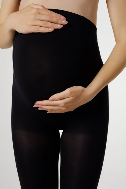 A pregnant woman wearing opaque black maternity tights that cover her belly and legs against a plain white background.