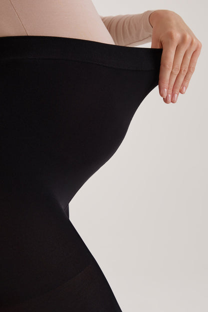 A pair of black maternity leggings being held out by a hand to show the fabric's stretch over a pregnant belly.