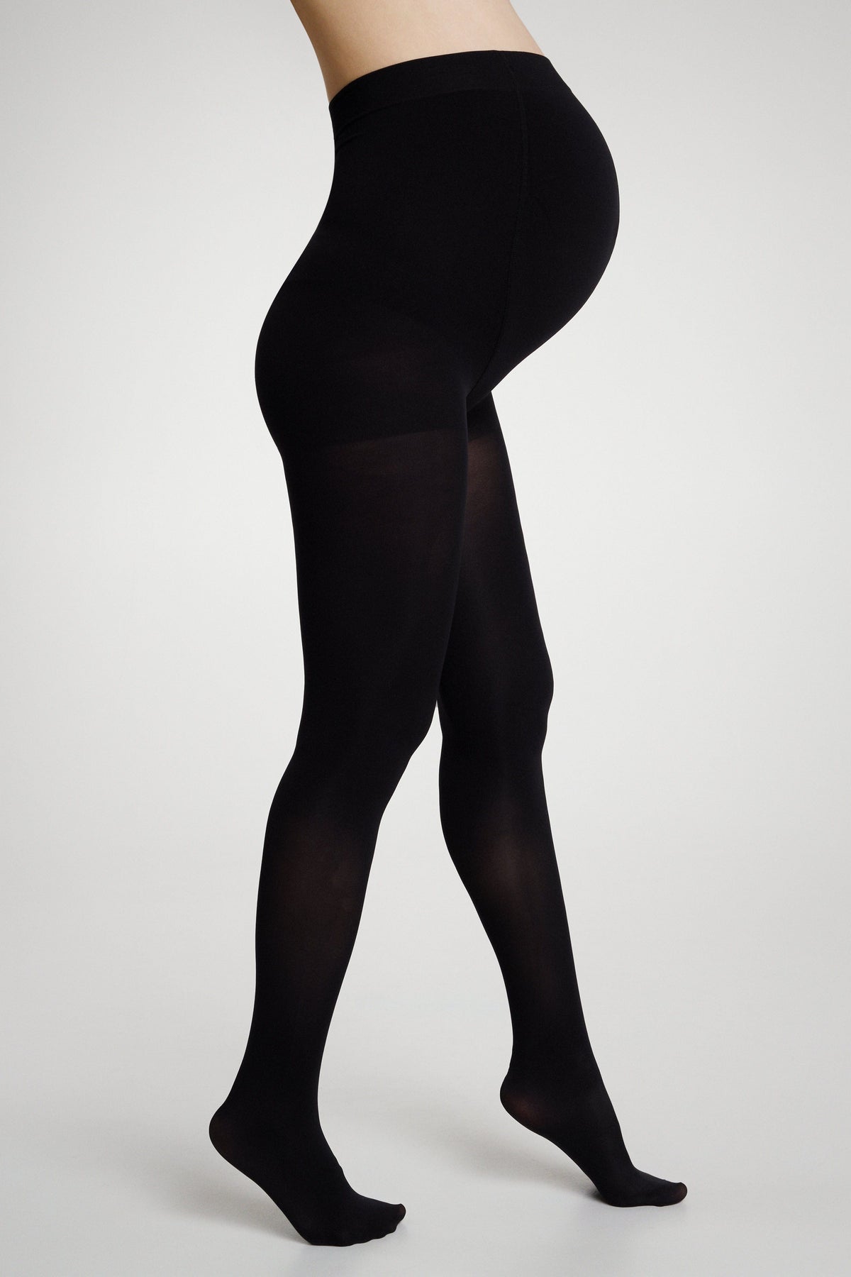 A pregnant woman wearing opaque black maternity tights stands in profile against a plain light gray background.