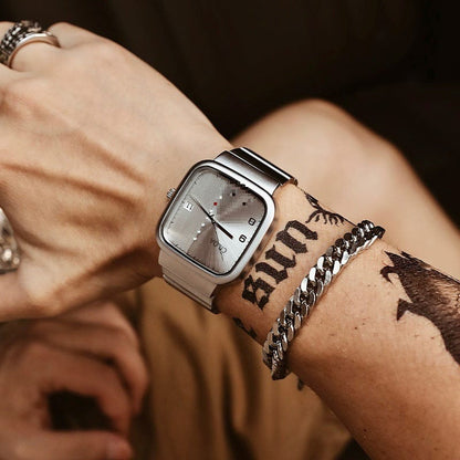A silver-toned square wristwatch with a metallic link strap worn on a tattooed wrist alongside a silver chain bracelet.