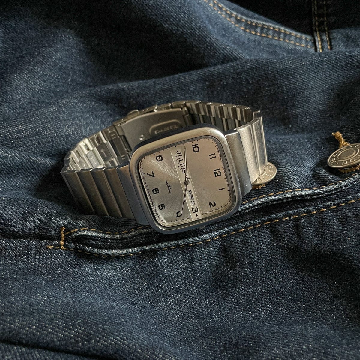 A silver-toned wristwatch with a square dial and a link bracelet rests on a dark denim fabric surface.