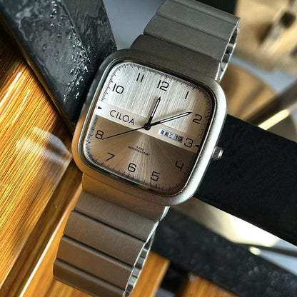 A rectangular CILOA watch with a silver brushed dial, black Arabic numerals, and a metallic bracelet band resting against a wooden surface.