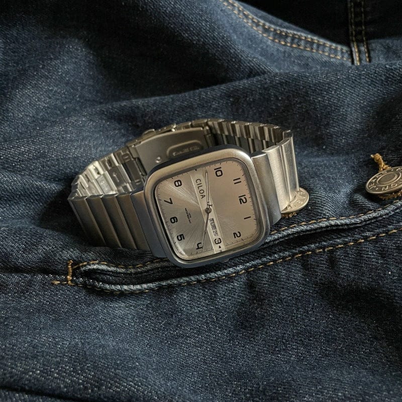 A silver-toned square wristwatch with a white dial, black numerals, and a day-date display rests on dark blue denim fabric.