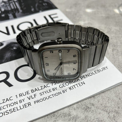 A silver-toned rectangular watch with a metallic bracelet band resting on a black and white print magazine.
