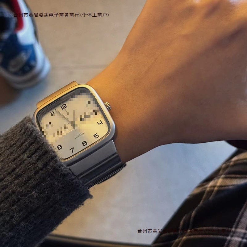 A silver-toned square-faced wristwatch with a white dial, Arabic numerals at 4, 5, 6, 7, 8, and 12, and a metal link strap worn on a person's wrist.