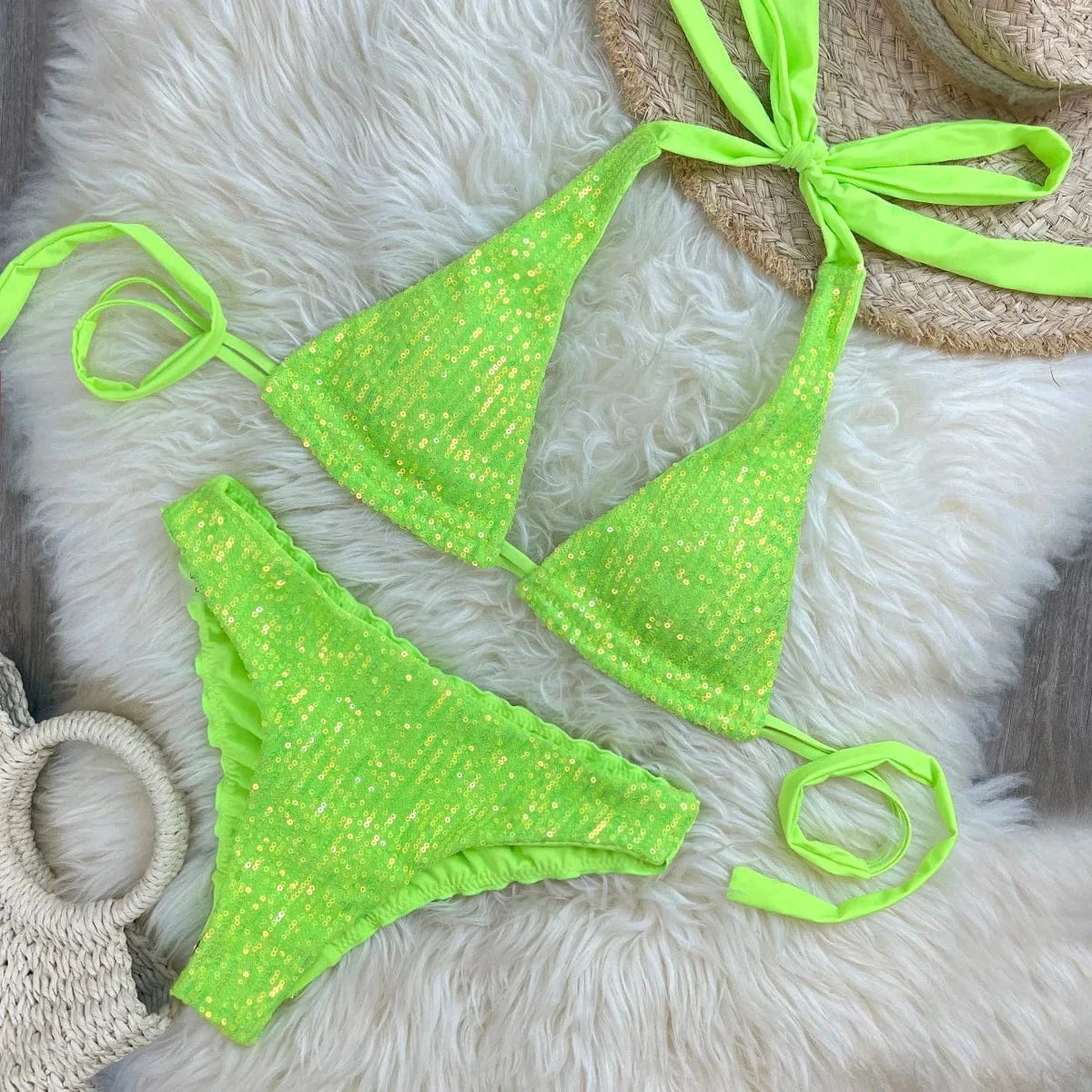 A bright neon green sequin bikini set consisting of a triangle top with tie-back straps and matching cheeky bottoms, displayed on a textured white faux-fur rug next to a straw hat and a woven bag.