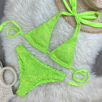 A bright neon green sequin bikini set consisting of a triangle top with tie-back straps and matching cheeky bottoms, displayed on a textured white faux-fur rug next to a straw hat and a woven bag.