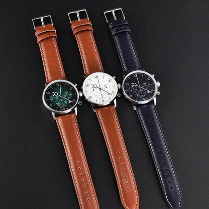 Three wristwatches with round silver cases and leather straps are arranged side-by-side on a dark background, featuring a green dial with a tan strap, a white dial with a tan strap, and a navy blue dial with a matching navy blue strap.