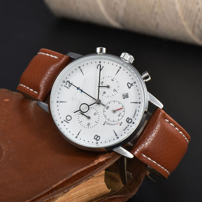 A round chronograph watch with a white dial, brown leather strap, and silver-tone case resting on a leather-bound book against a dark background.