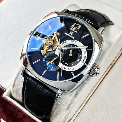 A silver-toned square wristwatch with a blue face, an open-heart dial, a moon phase display, and a black leather strap resting inside a white box.