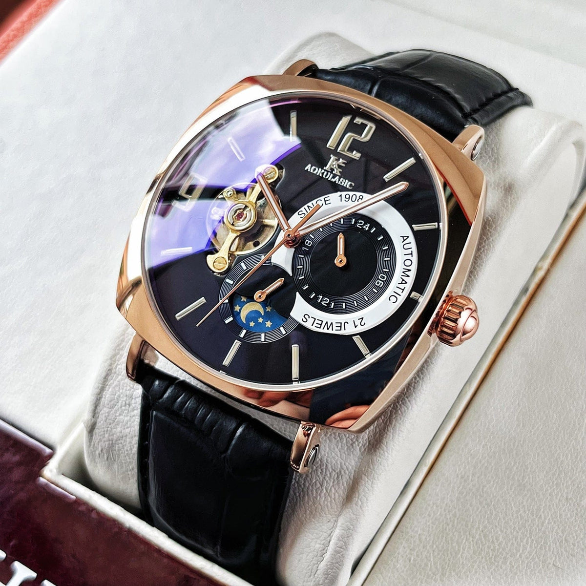 A rose gold square watch with a black dial, an open heart balance wheel, a moon phase display, and a black leather strap sits inside a white padded presentation box.