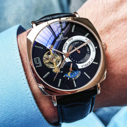 A rose gold-tone square-cased automatic watch with a dark blue dial featuring an open heart balance wheel, a day-night subdial, and a black leather strap worn on a wrist.