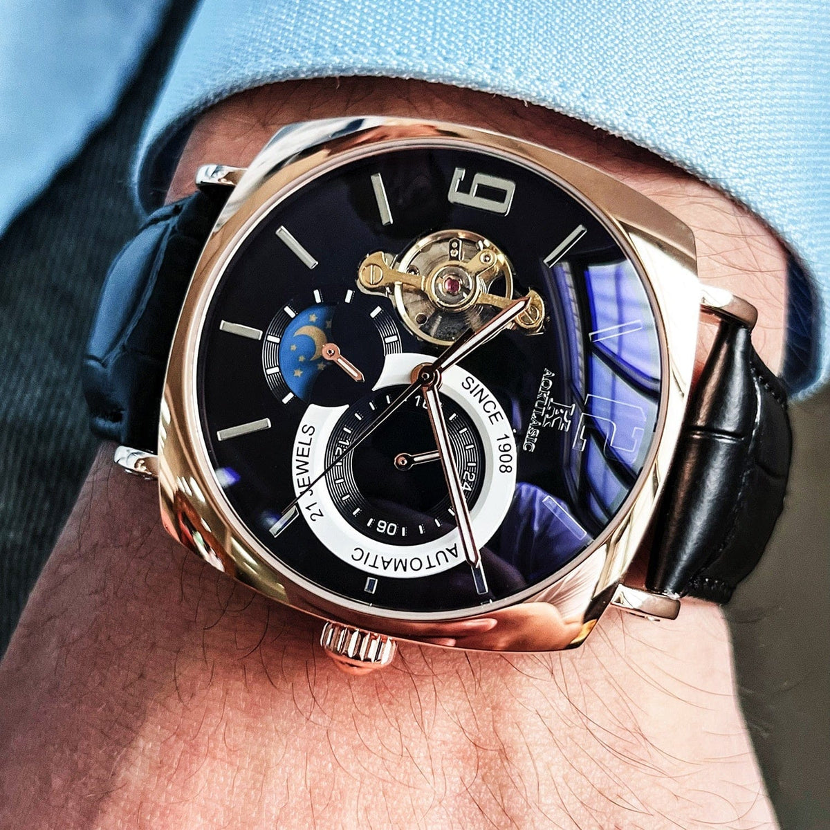 A close-up of a cushion-shaped wristwatch with a navy blue dial, rose gold accents, an exposed balance wheel, a moon phase sub-dial, and a black leather strap worn on a wrist.