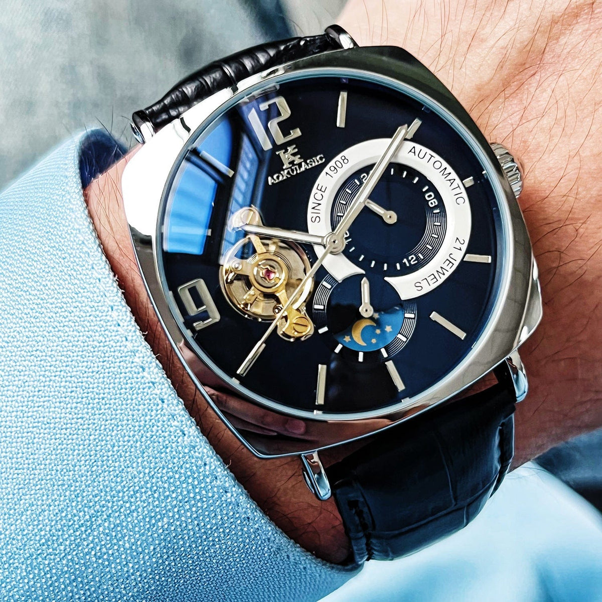 A close-up of a wrist wearing an analog wristwatch with a dark blue face, a silver-toned square-shaped case, and a black leather strap featuring a visible balance wheel and a moon phase display.