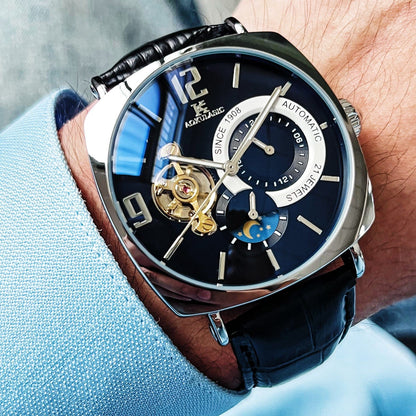 A close-up of a wrist wearing an analog wristwatch with a dark blue face, a silver-toned square-shaped case, and a black leather strap featuring a visible balance wheel and a moon phase display.
