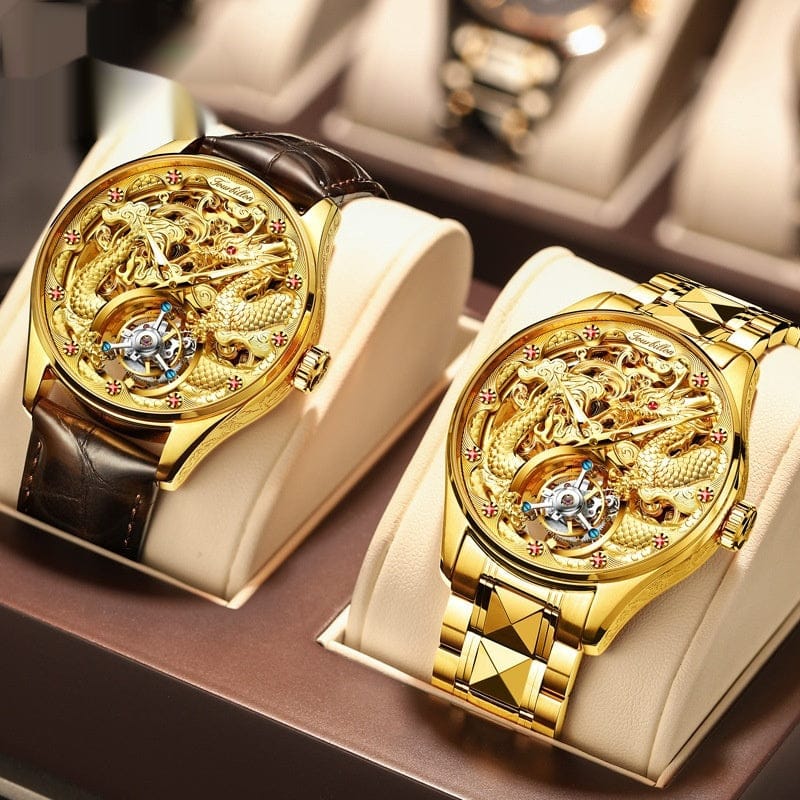 Two gold-toned skeleton watches featuring ornate dragon designs on the dials are displayed side-by-side on beige watch cushions inside a display box, with one watch on a brown leather strap and the other on a matching gold metal link bracel