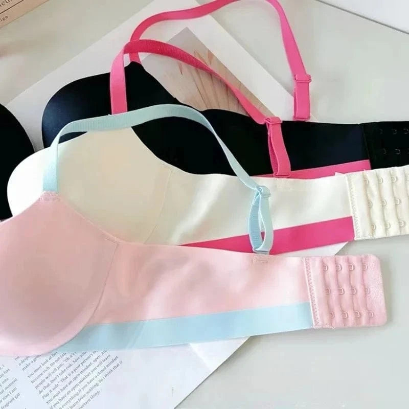 Three bras are stacked on top of each other, featuring smooth cups in black, white, and pale pink with contrasting light blue or pink adjustable straps and matching hook-and-eye back closures.